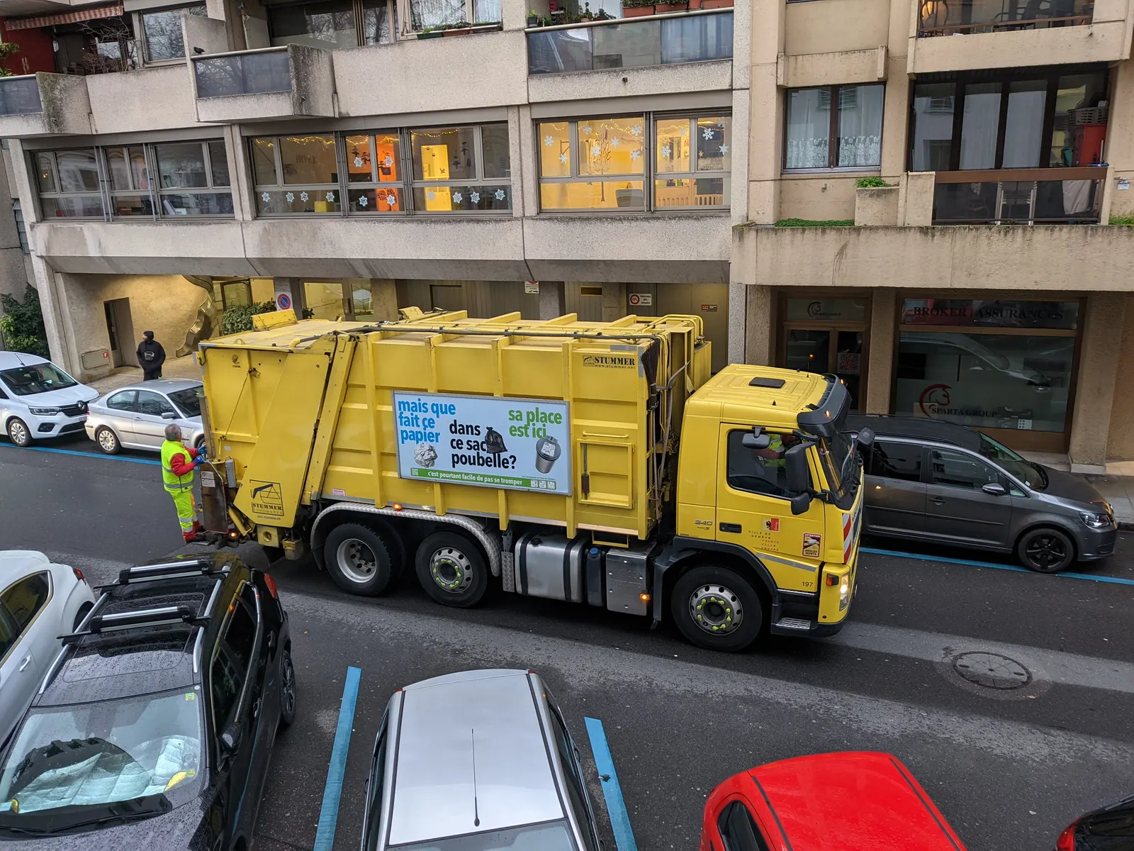 A yellow municipal refuse collection truck working a residential street in Geneva while a hi-vis crew member loads waste at the rear