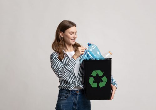 Waste Recycling Concept. Smiling Young Woman Holding Black Container With Plastic Bottles And Green Recycle Sign, Millennial Eco-Friendly Lady Standing Over Gray Background In Studio, Copy Space